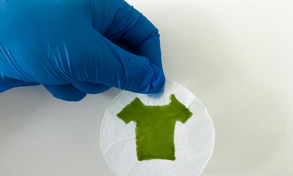 a blue-gloved hand holds a tiny circle of white material with a tiny green t-shirt printed on it.